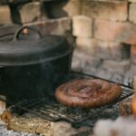 A covered pot sits on a hot grate next to a coiled sausage as they cook in a hearth