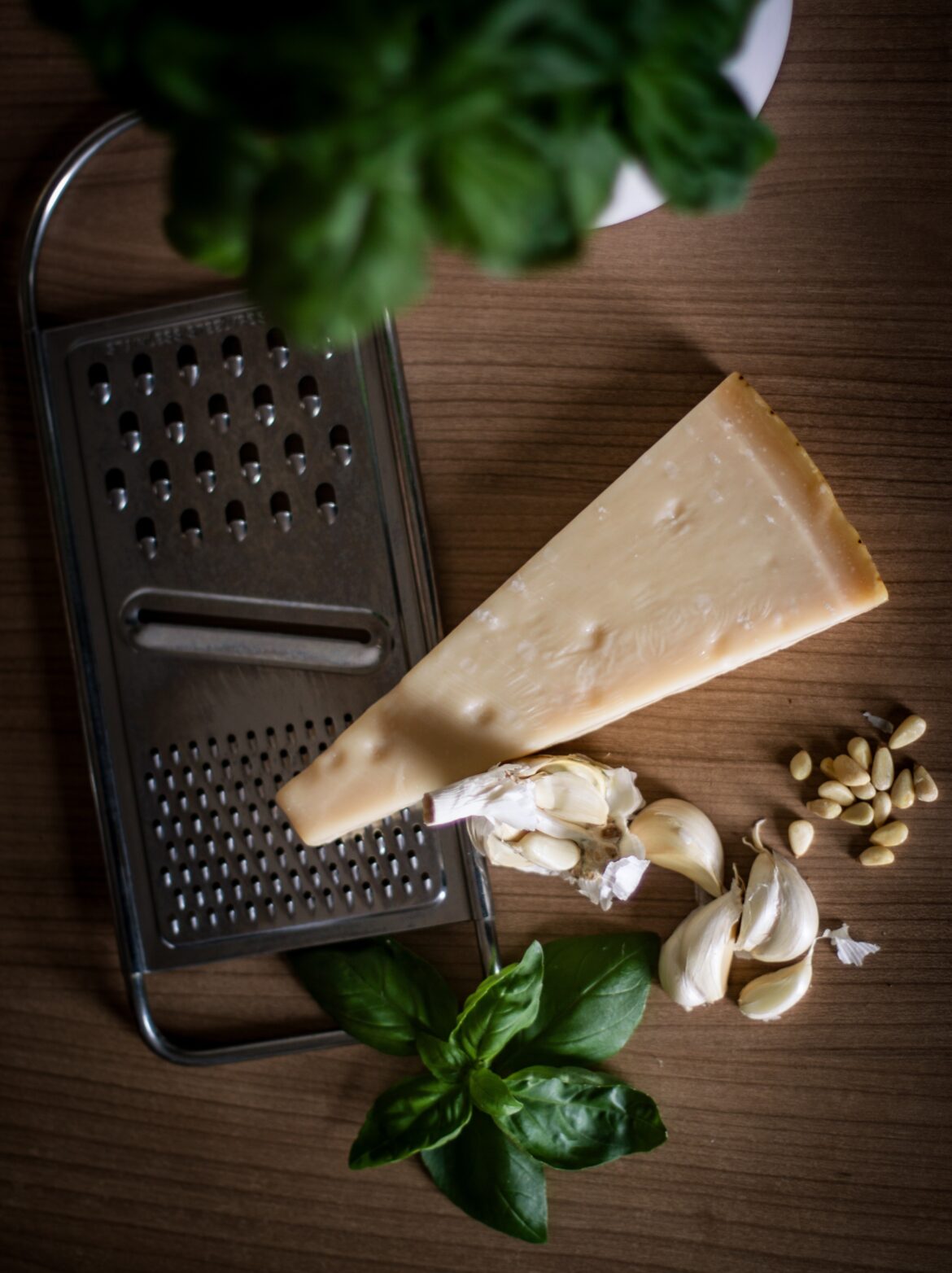A wedge of Parmesan cheese sits on top of a grater alongside cloves of garlic and fresh basil