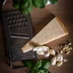 A wedge of Parmesan cheese sits on top of a grater alongside cloves of garlic and fresh basil